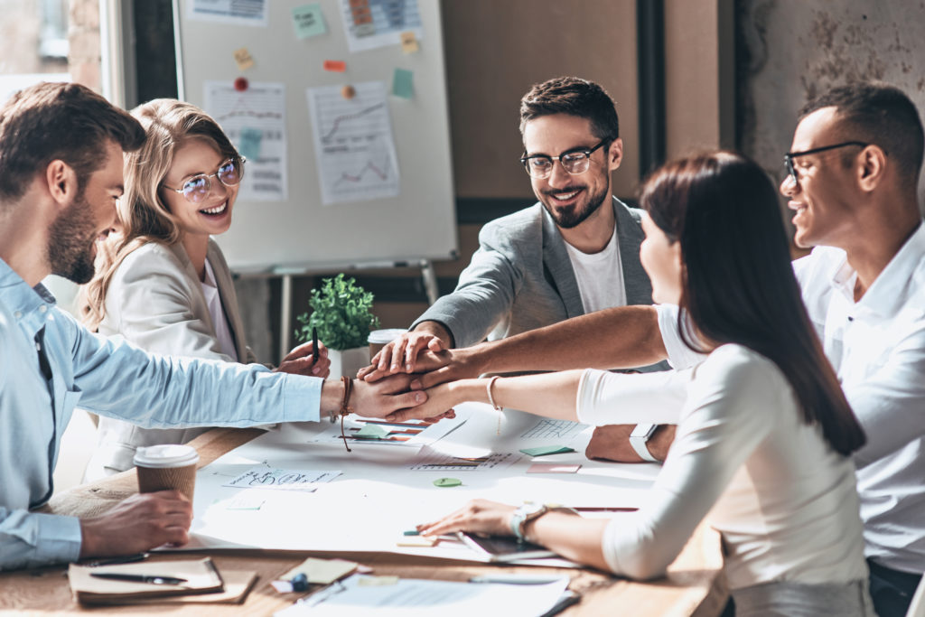 Teamwork. Young modern people in smart casual wear holding hands on top of one another in a symbol of unity while working in the creative office